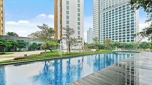 A modern swimming pool with a wooden deck, surrounded by greenery and tall buildings. - Anera by Kozystay - Cilandak (South Jakarta)