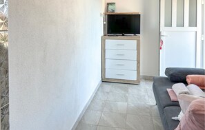 A living room with a flat-screen TV on a wooden stand, a grey sofa with a white cushion, and a tiled floor. - Lovely apartment in Rogoznica with WiFi (Rogoznica)
