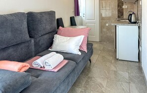 Ein graues Sofa mit aufgerollten Handtüchern und Kissen, eine weiße Tür und ein Küchenbereich mit einem Wasserkocher.