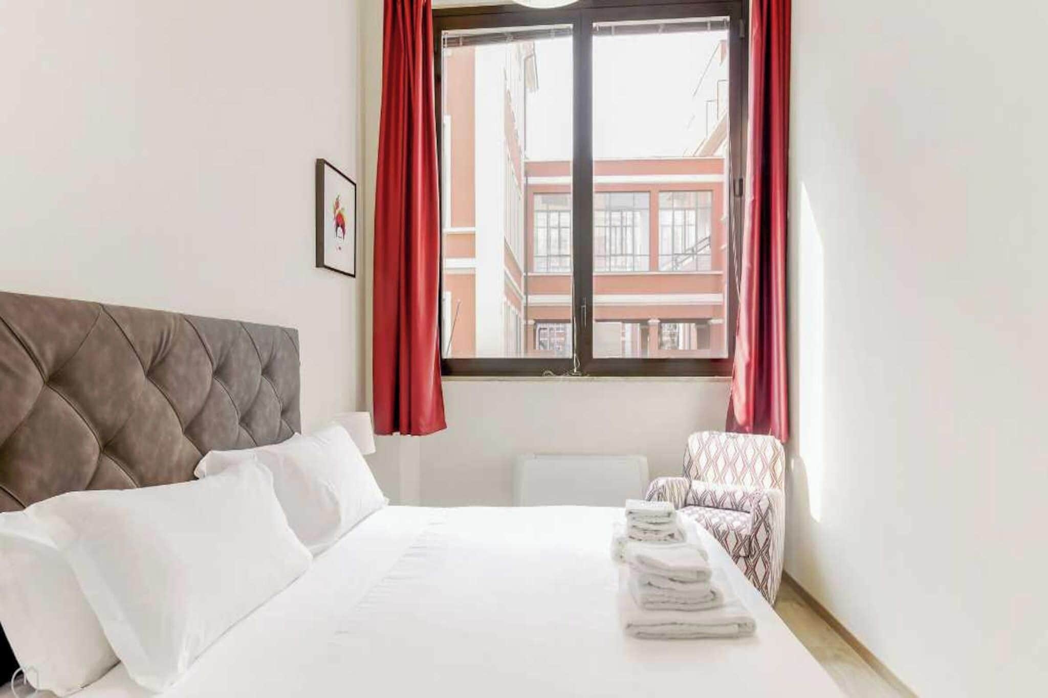 A hotel room with a bed, a chair, a window with curtains, and a framed picture on the wall.