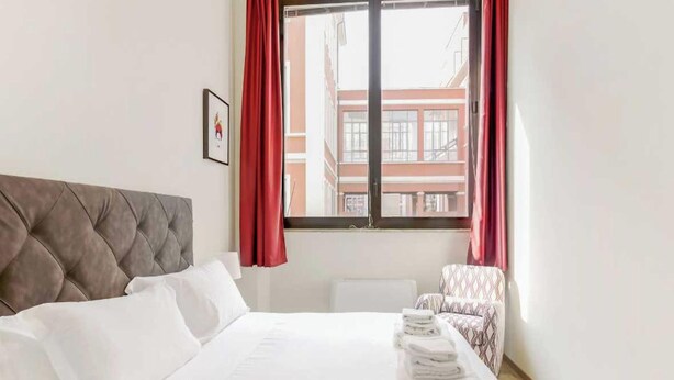Ein Hotelzimmer mit Bett, Stuhl, einem Fenster mit Vorhängen und einem gerahmten Bild an der Wand.