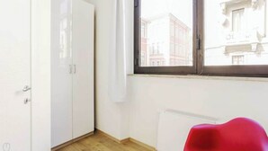 Ein Zimmer mit einem weißen Kleiderschrank, einem Fenster mit Blick auf Gebäude und einem roten Sessel.