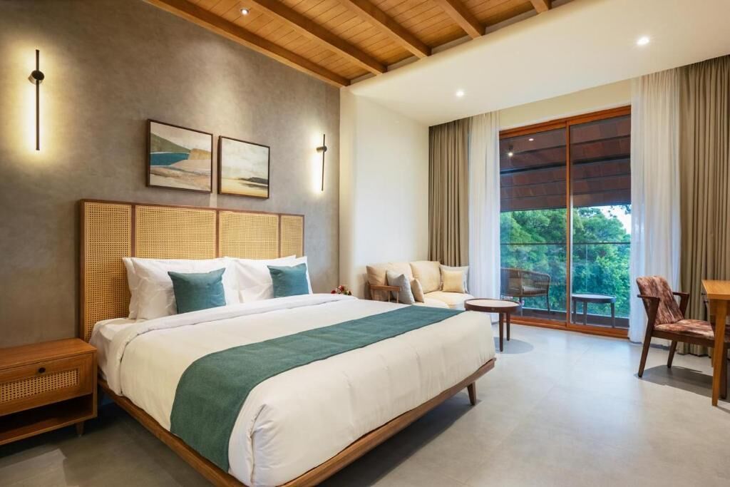 Habitación de hotel moderna con una cama grande, zona de estar con una mesa y vistas a zonas verdes por la ventana.