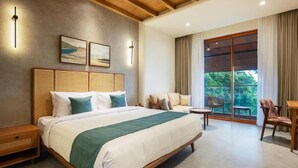 A modern hotel room with a large bed, a seating area with a table, and a view of greenery through the window. - Olive Woods (CHINNAKANAL)