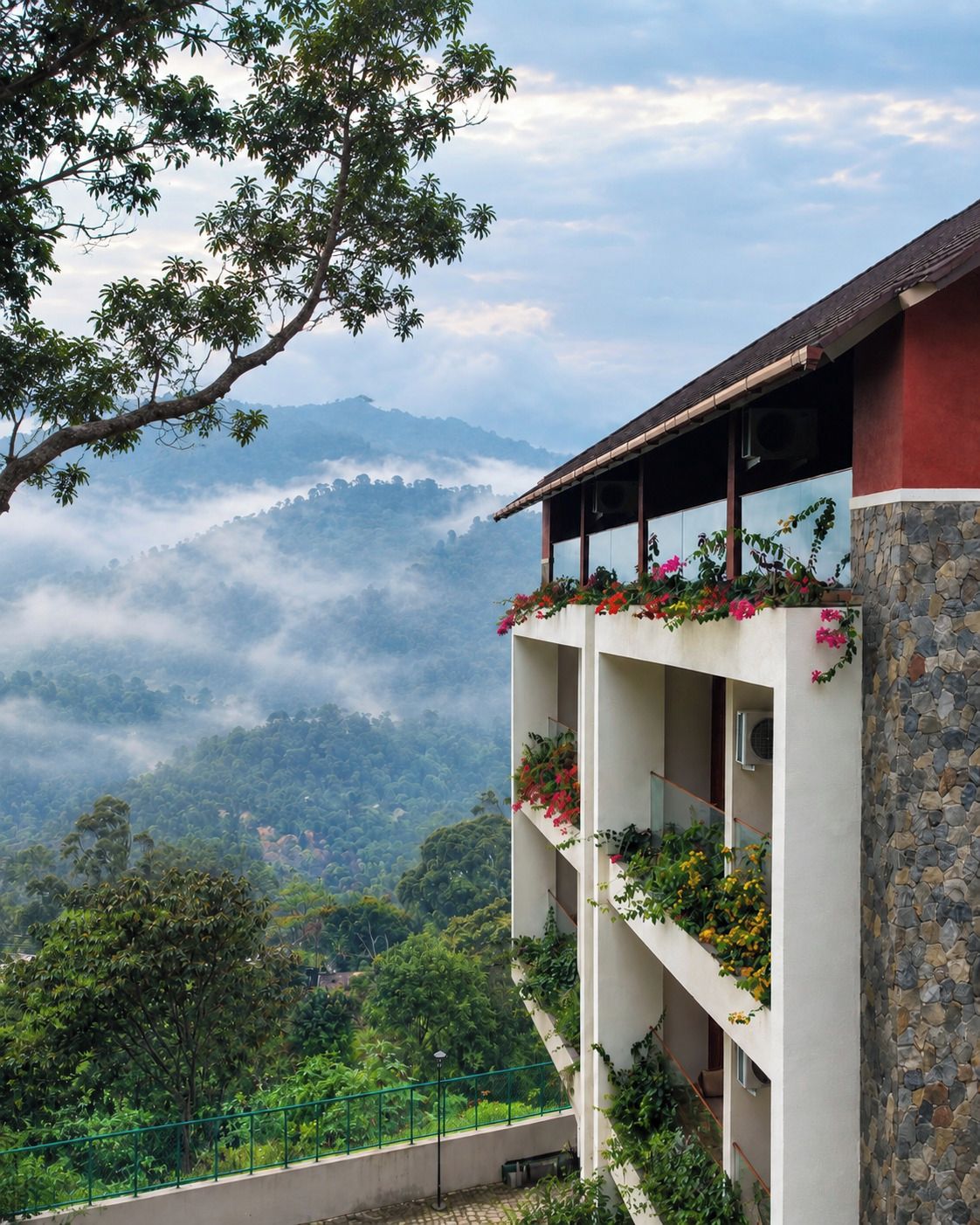 A multi-story building with balconies, surrounded by lush greenery and a mountainous backdrop.
