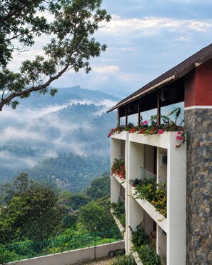 A multi-story building with balconies, surrounded by lush greenery and a mountainous backdrop. - Olive Woods (Devikolam)