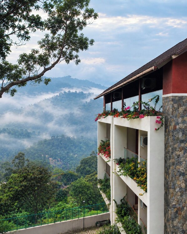 A multi-story building with balconies, surrounded by lush greenery and a mountainous backdrop. - Olive Woods (Devikolam)