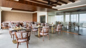 A spacious dining area with multiple tables and chairs, a modern ceiling design, and large windows offering a view. - Olive Woods (CHINNAKANAL)