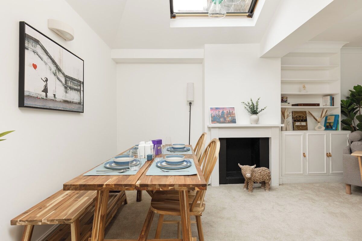 Uma sala de jantar com uma mesa de madeira posta para quatro pessoas, uma lareira branca e uma pequena ovelha decorativa.