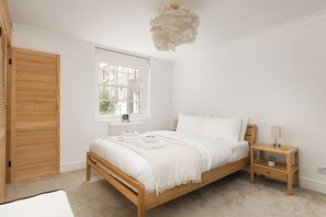 A bedroom with a wooden bed, a bedside table with a lamp, a window with blinds, and a radiator. - New! Bright 2BR Duplex w/Patio in Pimlico (Greater London)