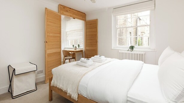 A bedroom with a bed, a wooden wardrobe, a small table, a chair, and a window with a view of buildings. - New! Bright 2BR Duplex w/Patio in Pimlico (Greater London)