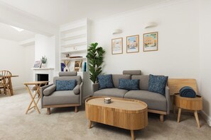 Ein modernes Wohnzimmer mit einer Couch, Sesseln, einem Couchtisch und einem Beistelltisch. Ein Kamin, eine Pflanze und gerahmte Bilder schmücken die Wand.