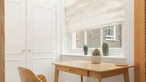 A room with a wooden desk, a chair, a window with a view of a brick wall and cacti, and a white door. - New! Bright 2BR Duplex w/Patio in Pimlico (Greater London)
