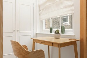 Ein Zimmer mit einem Holztisch, einem Stuhl, einem Fenster mit Blick auf eine Ziegelwand und Kakteen sowie einer weißen Tür.