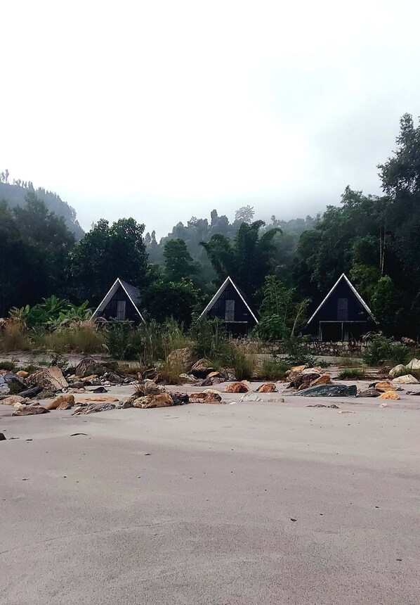 Three triangular-roofed cabins with a stone wall in front, set in a natural landscape with trees and a road in the foreground.
