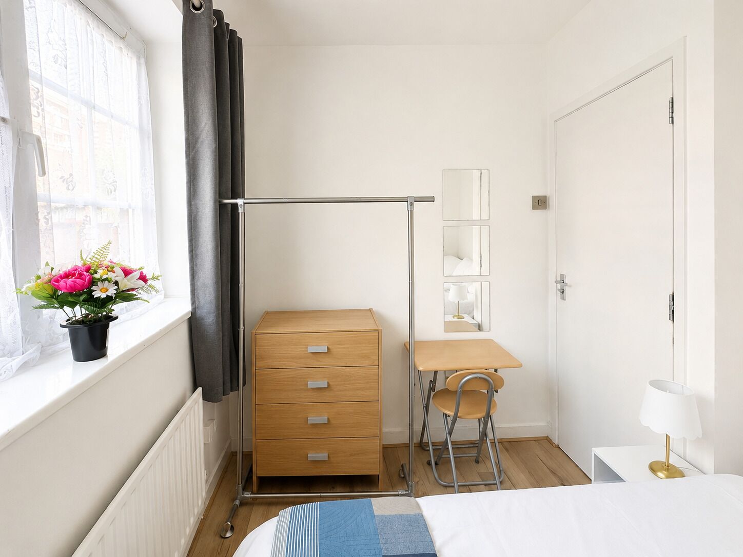 Un dormitorio con cama, cómoda de madera, escritorio con silla, radiador, ventana con cortinas y planta en maceta con flores.
