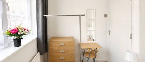 Une chambre à coucher comprenant un lit, une commode en bois, un bureau avec une chaise, un radiateur, une fenêtre avec des rideaux et un pot de fleurs.