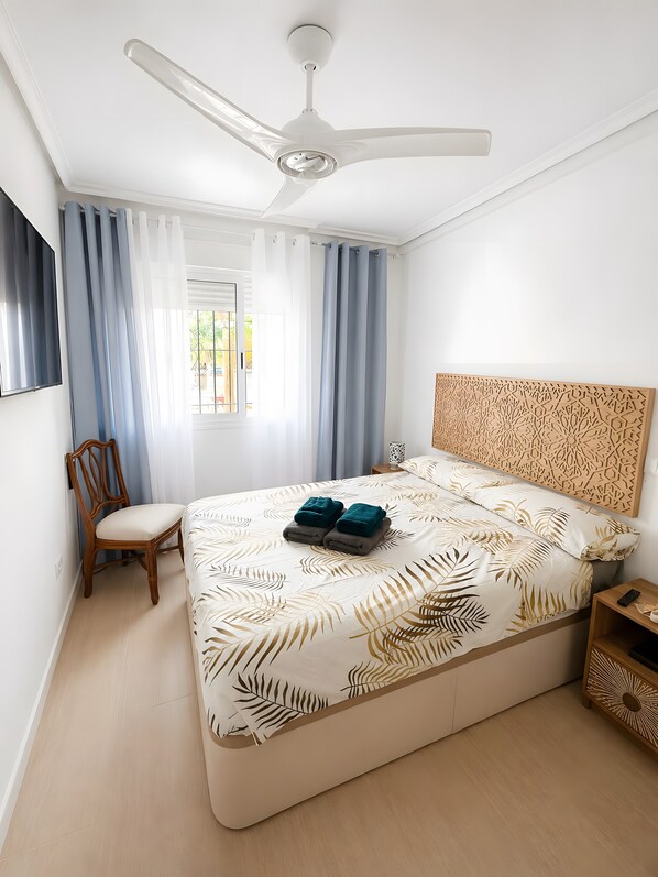 A bedroom with a bed, a ceiling fan, a chair, a nightstand, and a window with curtains.
