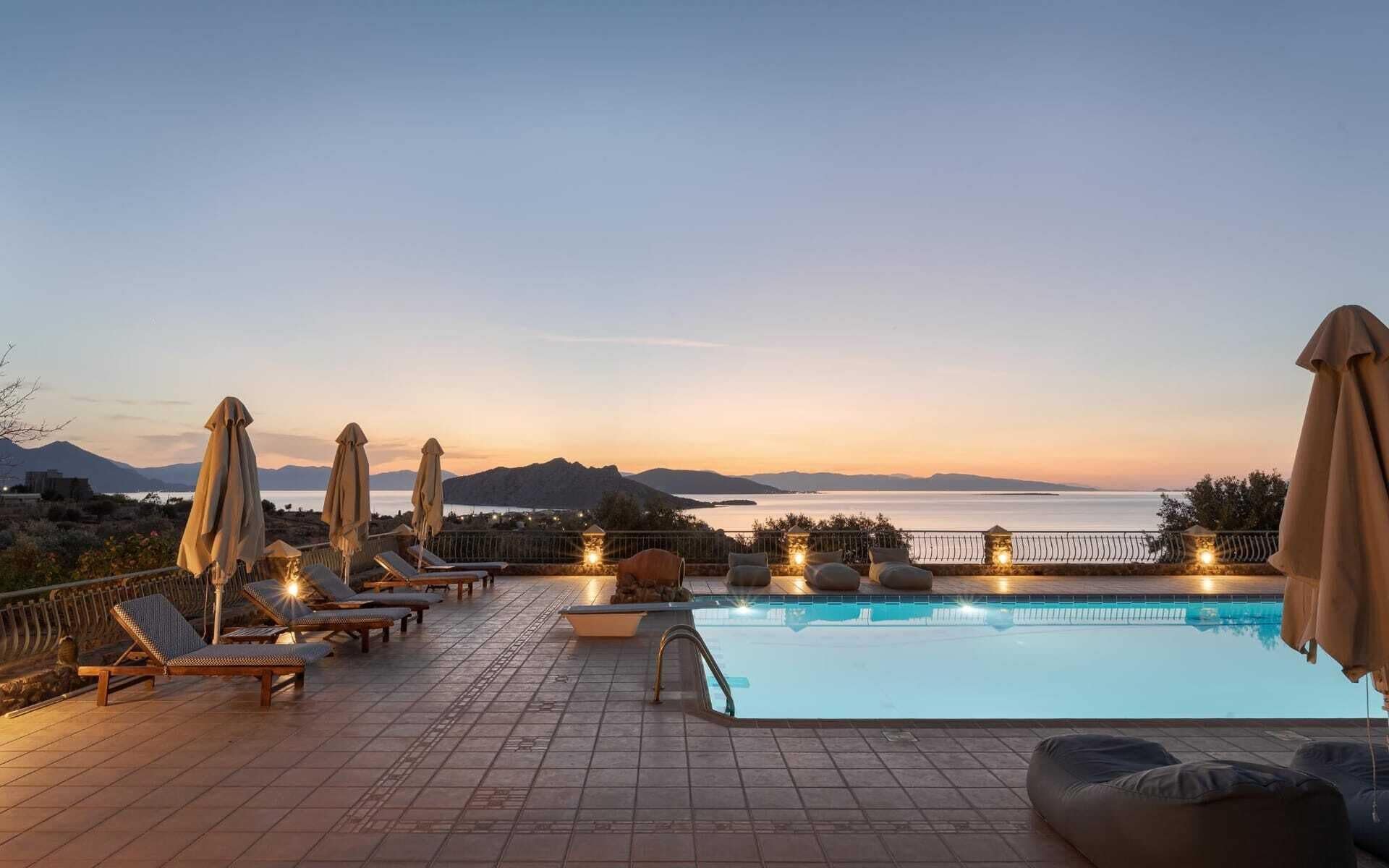 A poolside area with lounge chairs and umbrellas, overlooking a body of water and mountains.