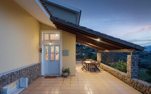 A covered patio with a dining table and chairs, a stone wall, and a door with a light fixture. - Aelia Villa Aegina, Villa (Aegina)