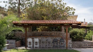 A stone-built outdoor bar area with a tiled roof, surrounded by greenery and a stone wall. - Aelia Villa Aegina, Villa (Aegina)