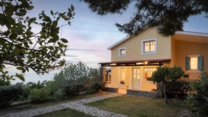 A two-story house with a stone pathway leading to the entrance, surrounded by greenery and a view of the sea. - Aelia Villa Aegina, Villa (Aegina)