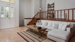 A living room with a white sofa, a wooden coffee table, a striped rug, a wooden staircase with a railing, and a bookshelf. - Aelia Villa Aegina, Villa (Aegina)