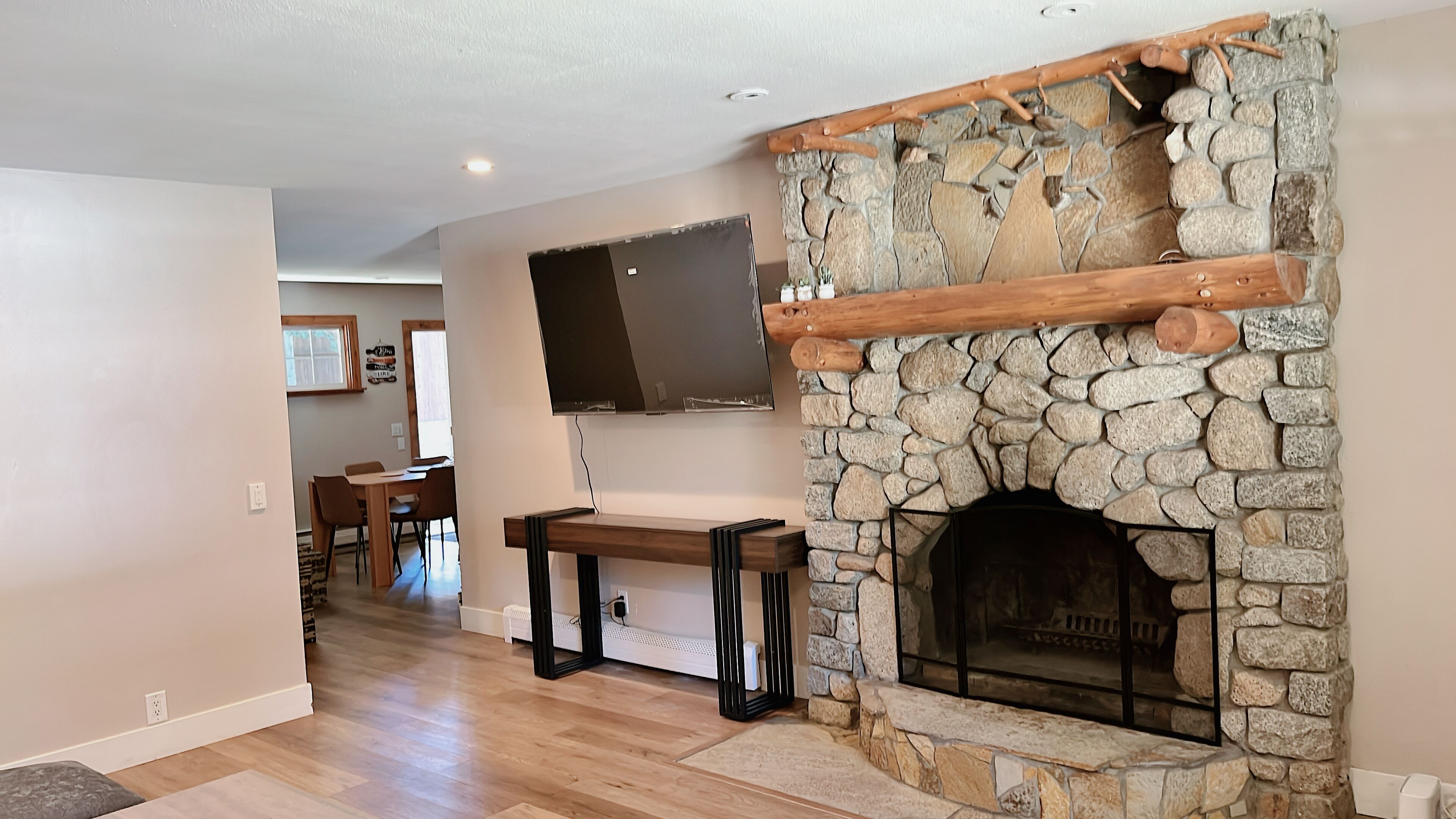 Una sala de estar con chimenea de piedra, televisor montado y un comedor al fondo.