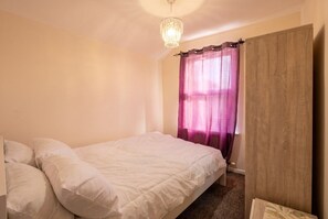 A bedroom with a bed, a chandelier, a window with curtains, and a wooden door. - 3-Bed Townhouse Sleeps 5 · Walk to Designer Outlet & STEAM Museum (Swindon)