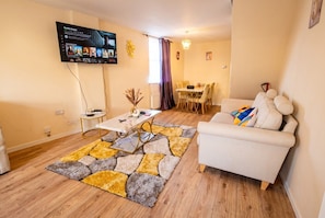 A living room with a sofa, a coffee table, a TV mounted on the wall, and a dining area in the background. - 3-Bed Townhouse Sleeps 5 · Walk to Designer Outlet & STEAM Museum (Swindon)