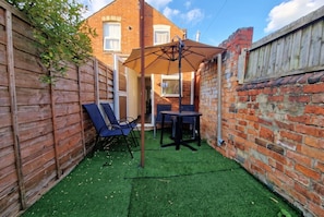 A backyard with artificial grass, a wooden fence, a brick wall, and a table with chairs. - 3-Bed Townhouse Sleeps 5 · Walk to Designer Outlet & STEAM Museum (Swindon)