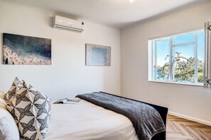 A bedroom with a bed, a window with a view, and an air conditioning unit on the wall. - Camps Bay 2 Bed with Sea Views Shoreline Views (Cape Town)