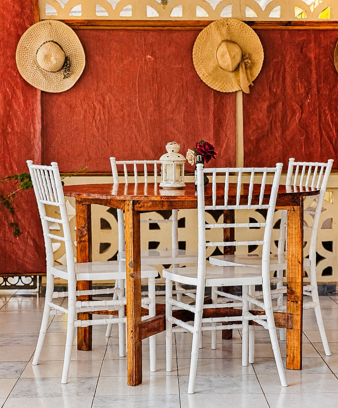 Un comedor con una mesa de madera y sillas blancas, un farol y dos sombreros en la pared.