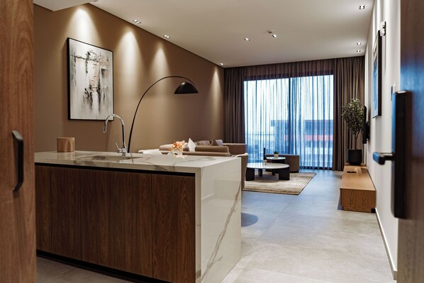 A modern kitchen with a marble countertop and wooden cabinets, leading to a living area with a sofa and a coffee table.