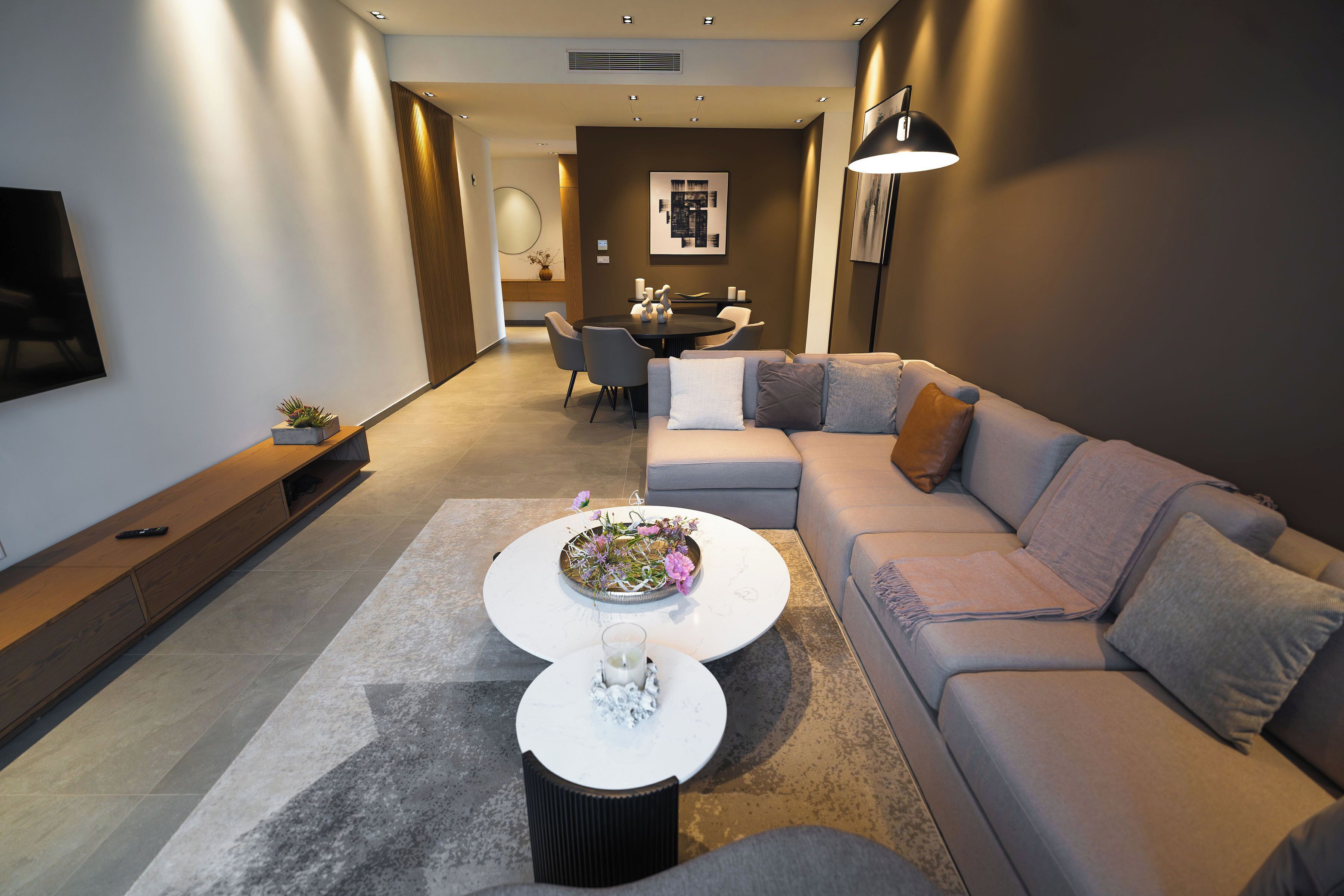 Una sala moderna con un sofá gris, una mesa de centro con flores, un televisor en la pared y un comedor al fondo.