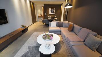 Una sala moderna con un sofá gris, una mesa de centro con flores, un televisor en la pared y un comedor al fondo.
