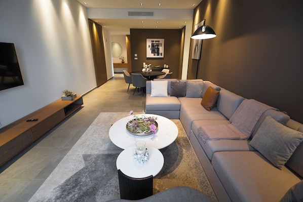A modern living room with a grey sofa, a coffee table with flowers, a TV on the wall, and a dining area in the background. - ICON RESIDENCES Douala (Douala)
