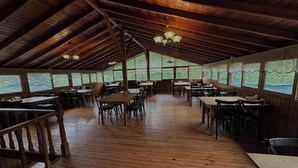 Un área de comedor cubierta con mesas y sillas de madera, ventanas amplias a lo largo de las paredes y un techo alto con vigas a la vista.