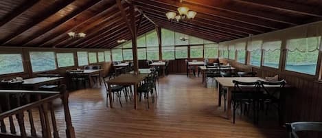 Un área de comedor cubierta con mesas y sillas de madera, ventanas amplias a lo largo de las paredes y un techo alto con vigas a la vista.
