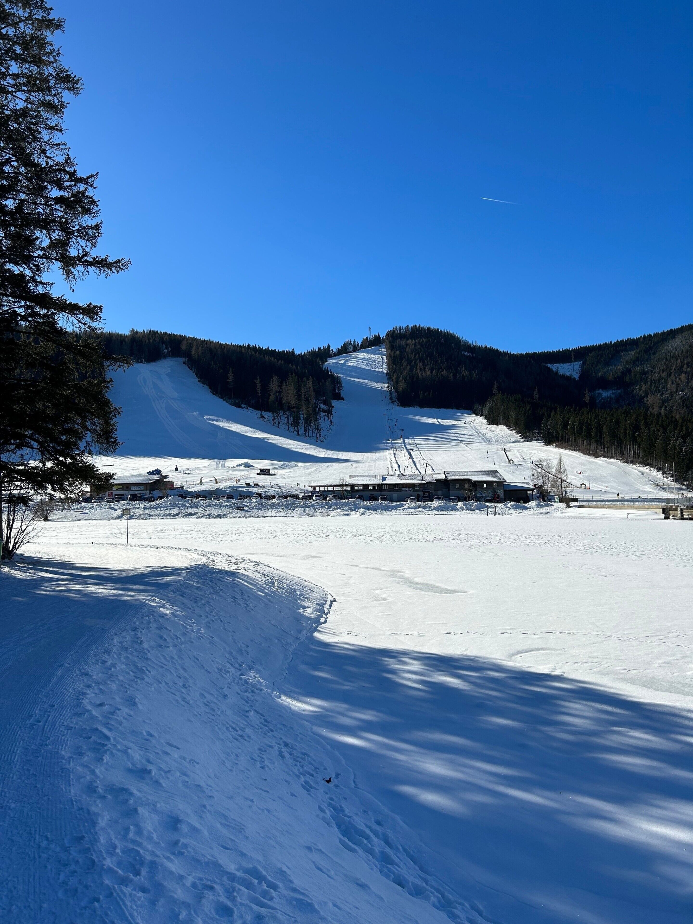 Pista de esqui coberta de neve, com céu claro, árvores e edifícios ao fundo.