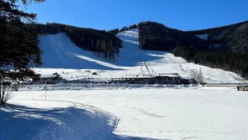 Pista de esqui coberta de neve, com céu claro, árvores e edifícios ao fundo.