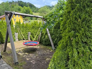 A wooden swing set with a colorful swing seat in a garden with dense shrubbery and a yellow building in the background.