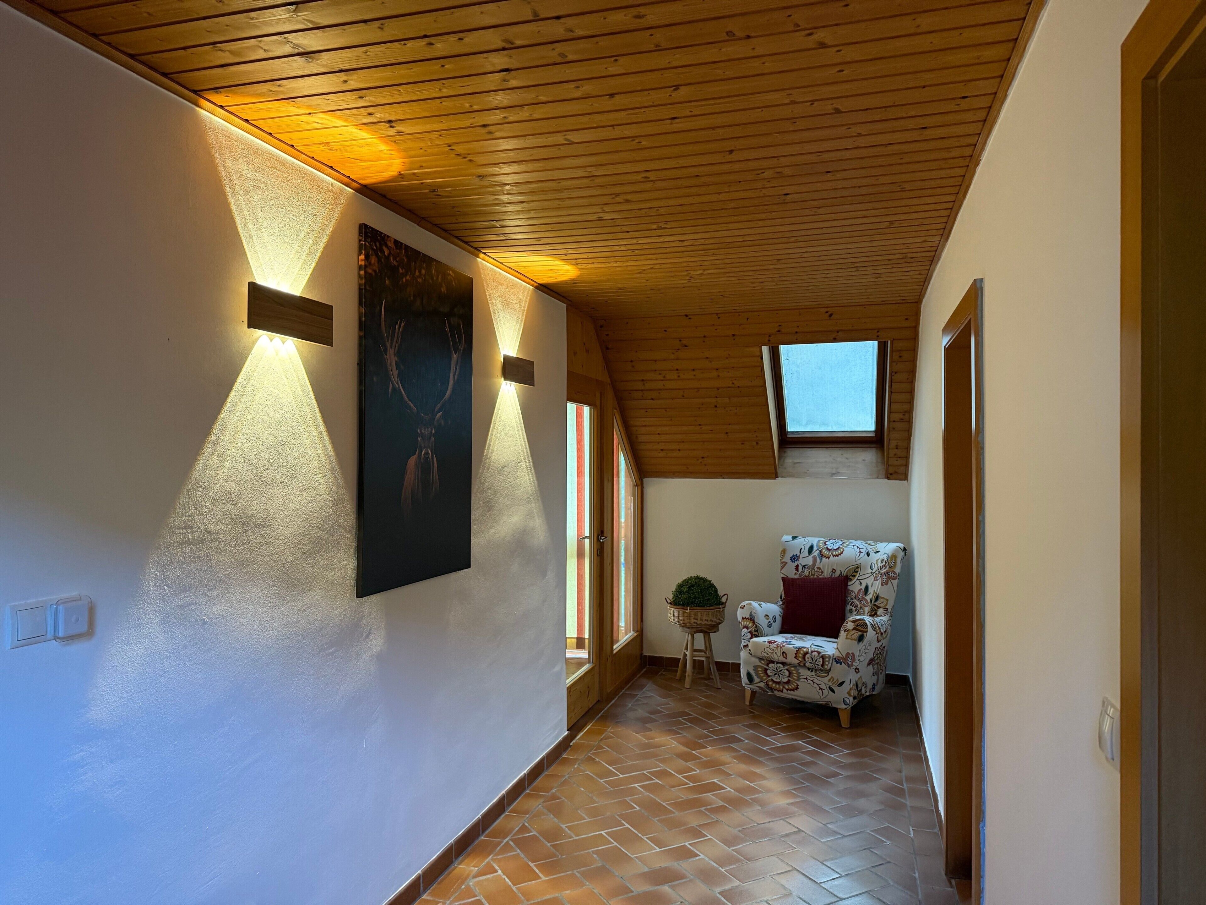 A hallway with a wooden ceiling, a patterned chair, a wall-mounted picture, and a small window.
