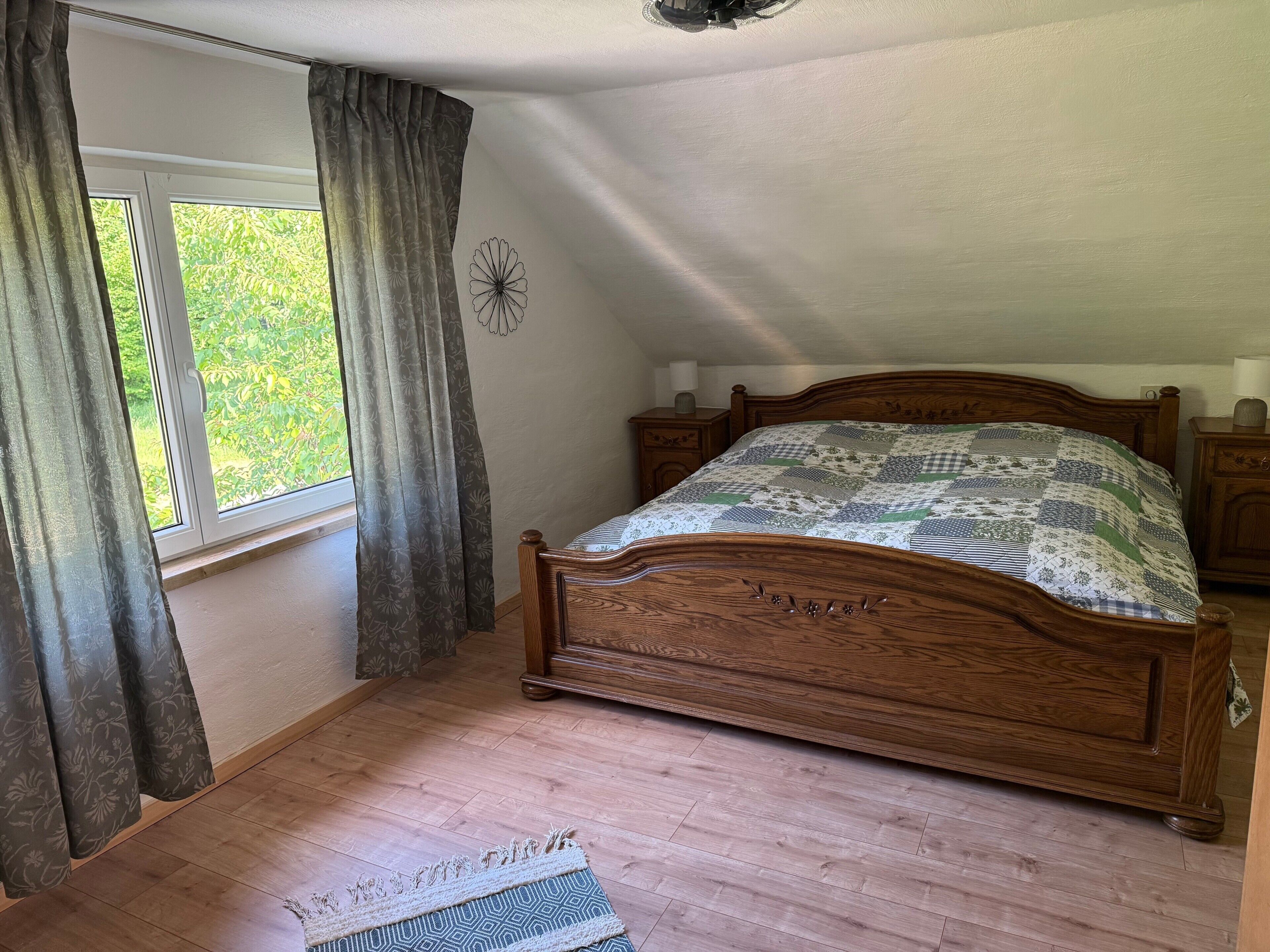 A bedroom with a wooden bed, a nightstand, a bedside table, a window with curtains, and a view of greenery outside.