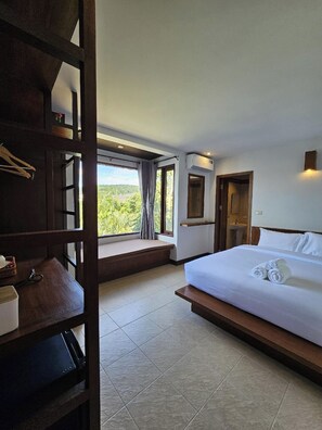 A hotel room with a large bed, a window with a view of greenery, a built-in shelf, and a bathroom with a sink and mirror. - Anda See House (Krabi)