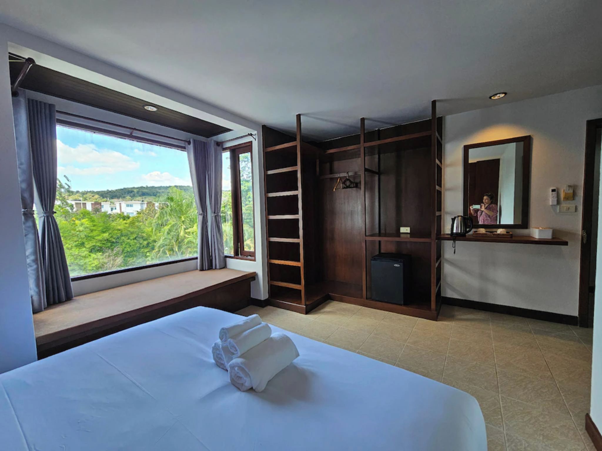 Une chambre d’hôtel avec un grand lit, une armoire, un petit réfrigérateur et une fenêtre donnant sur un paysage verdoyant.