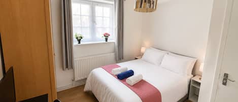 Een hotelkamer met een bed, een bureau, een stoel, een raam met gordijnen, een radiator en een hanglamp.