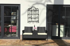 A black wicker bench with white cushions, a wall-mounted decorative panel with animal silhouettes, and a glass door with a reflection of a red table and chairs. - My Little Zanzibar 2 (Zirchow)