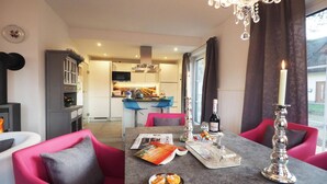 Un comedor moderno con una mesa dispuesta para dos personas, una araña y vista de la cocina al fondo.