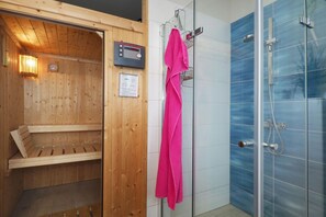 A sauna with wooden walls and a control panel, a glass shower enclosure with a blue-tiled wall, and a pink towel hanging on the shower door. - My Little Zanzibar 1 (Zirchow)